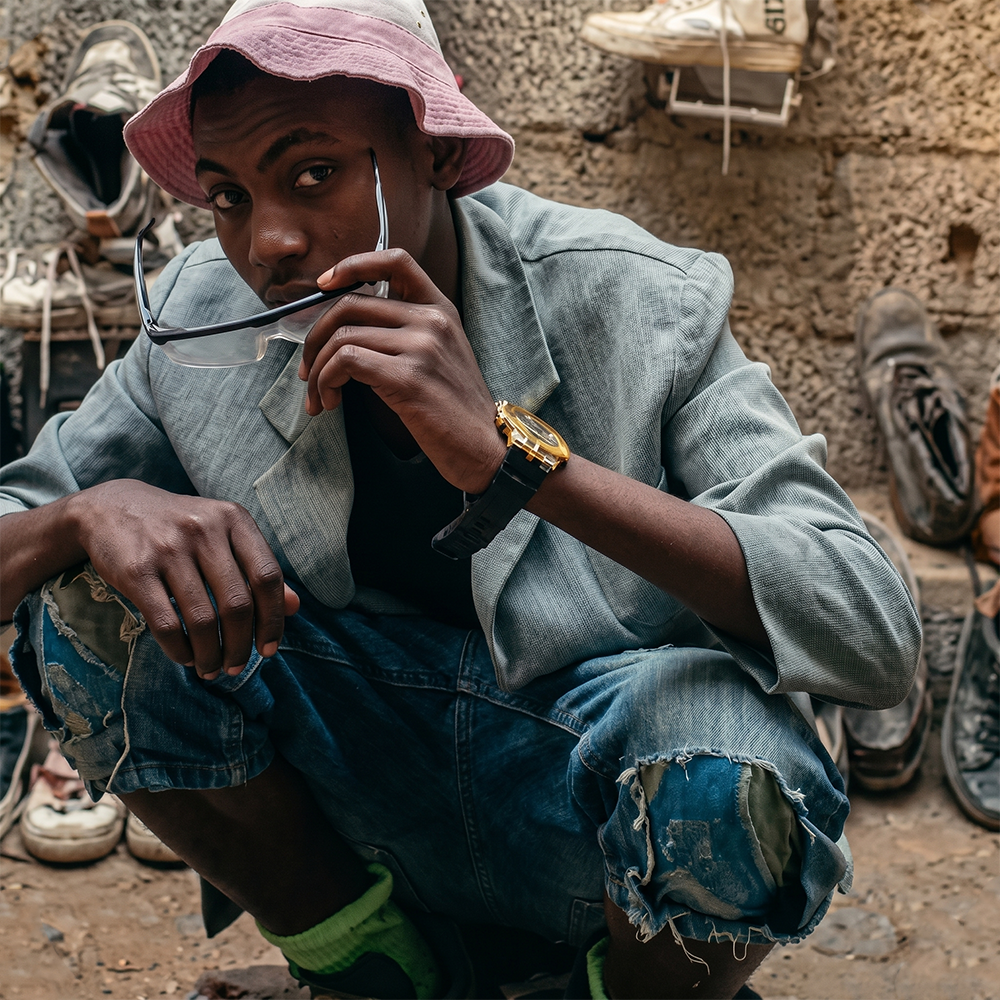 Working portrait of Kaluputics, crouched in denim and a pink bucket hat against a workshop wall.